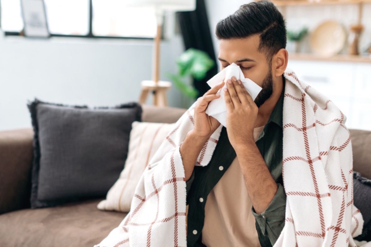 Person blowing their nose sits on the couch wrapped in a blanket